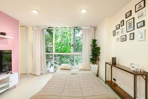 Ein Schlafzimmer mit großem Fenster, einem Bett, einem Fernseher, einer Pflanze und einer Wand mit gerahmten Bildern.