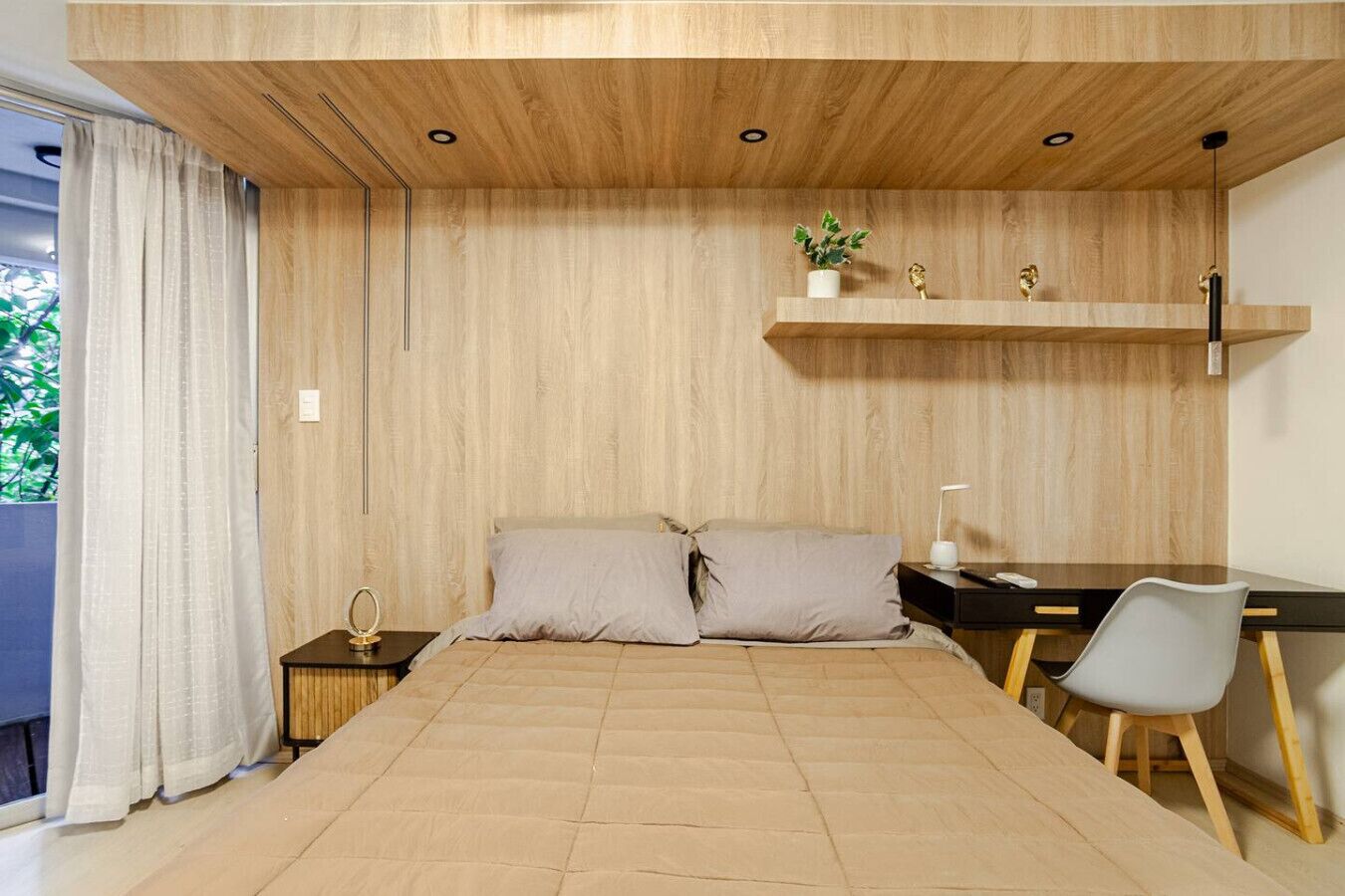 Una camera da letto moderna con un letto, una scrivania e una sedia. Si vede una finestra con tende, una mensola con oggetti decorativi e un tavolino con una lampada.