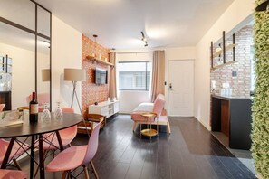A modern hotel room with a brick accent wall, a dining area with pink chairs, a small kitchen area, and a large window providing natural light.