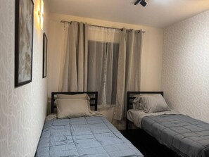 Quarto pequeno e bem organizado com duas camas de solteiro, uma janela com cortinas leves e um ponto de luz fixado na parede.