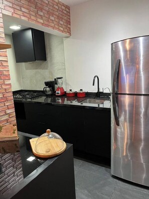 Uma cozinha moderna com geladeira de aço inoxidável, bancada preta, liquidificador vermelho e tábua de corte de madeira.