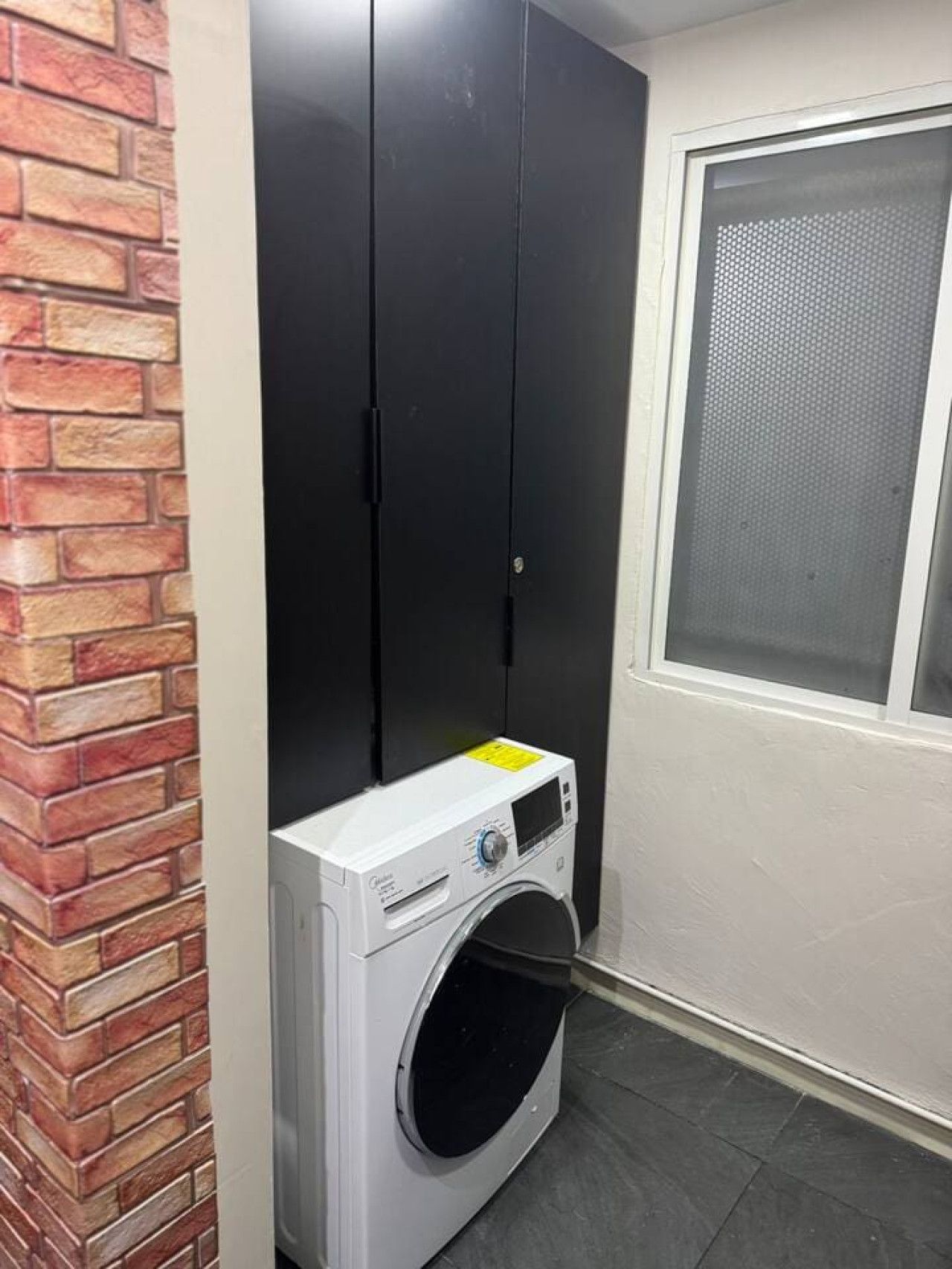 Een wasserette met een wasmachine, een zwarte garderobe en een raam met hor.