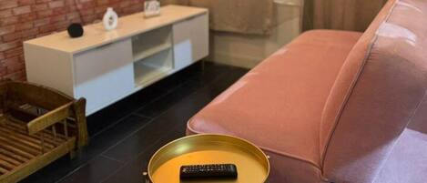 A modern living room with a TV mounted on a brick wall, a pink sofa, a small round table with a remote, and a wooden chair.