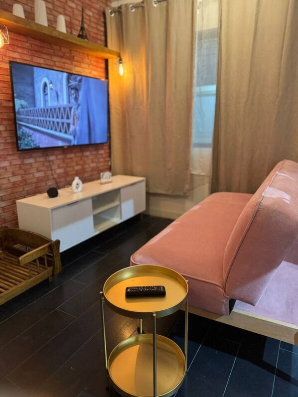 A modern living room with a TV mounted on a brick wall, a pink sofa, a small round table with a remote, and a wooden chair.