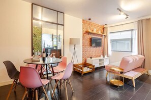 A modern living room with a round dining table, pink chairs, a brick accent wall, a TV mounted above a fireplace, and a large window with curtains.