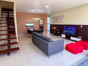 A modern living room with a grey sofa, red pouf, and a TV on a wooden stand.