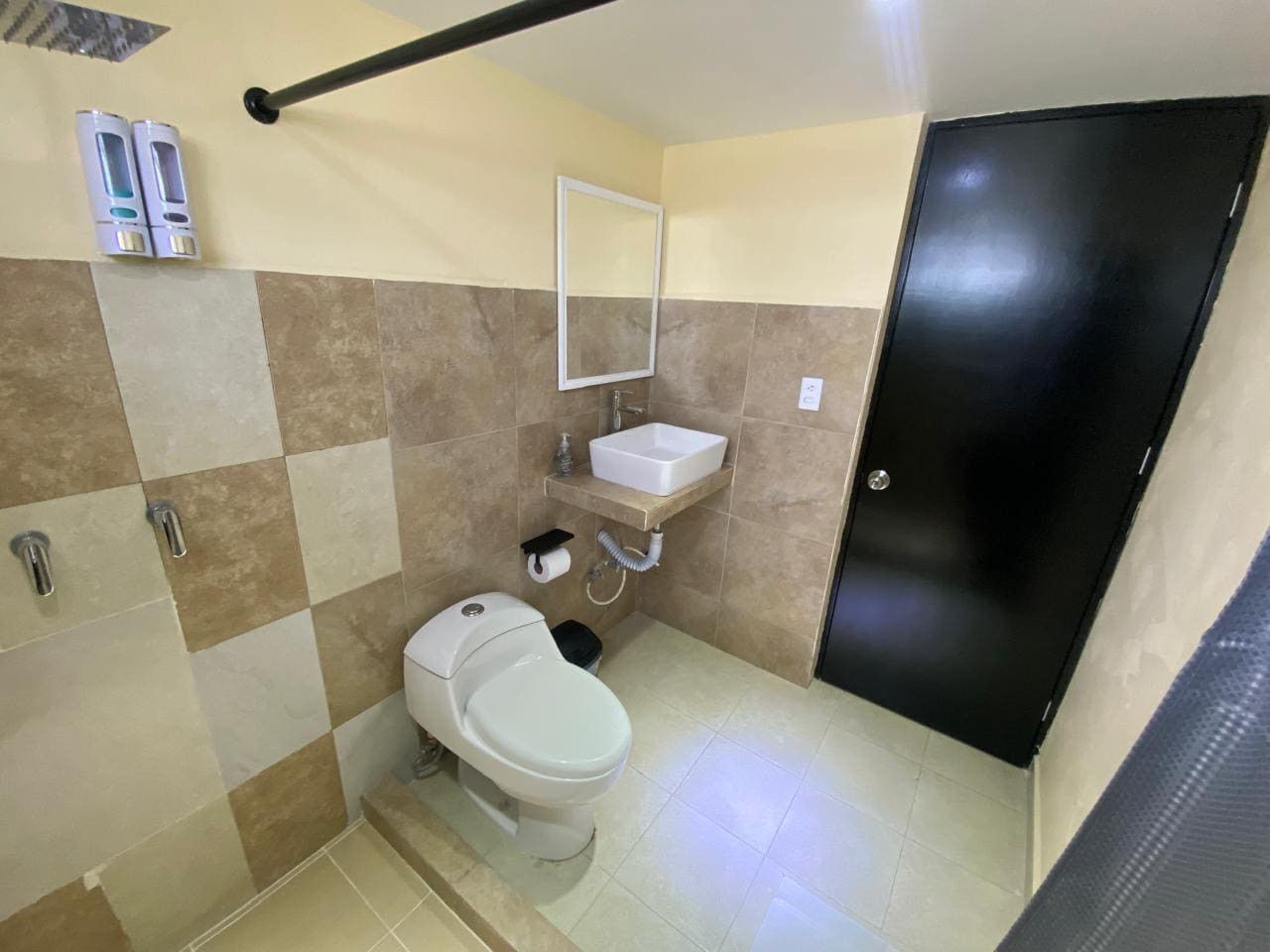 A small bathroom with a toilet, sink, and mirror. There are two soap dispensers on the wall, and a black door is visible.
