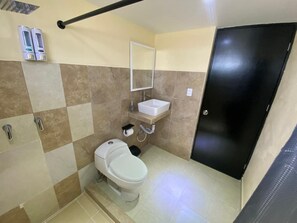 Une petite salle de bain avec des toilettes, un lavabo et un miroir. On peut voir deux distributeurs de savon fixés au mur, et une porte noire est visible.
