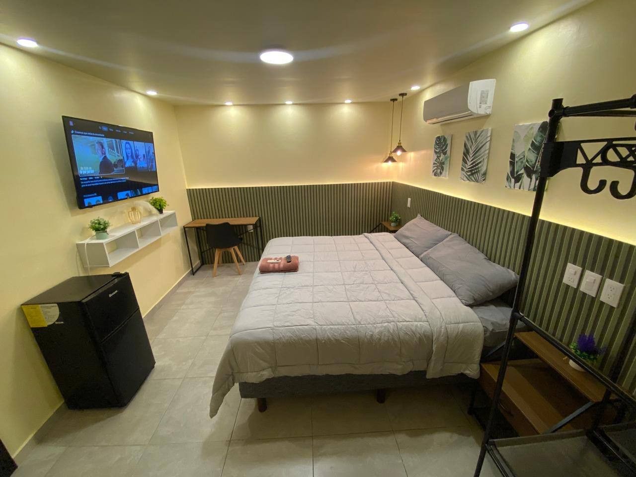 A compact hotel room with a bed, a desk, a chair, a TV, and a small shelf with decorative items.