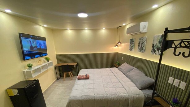 Habitación de hotel pequeña con cama, escritorio, silla, televisión y un estante con objetos decorativos.