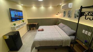 A compact hotel room with a bed, a desk, a chair, a TV, and a small shelf with decorative items.