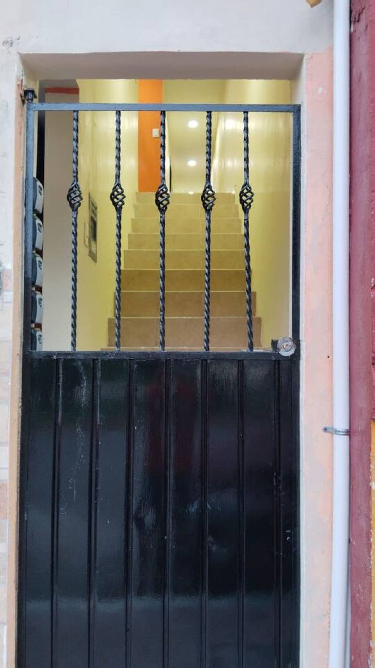 Una reja de metal negro con un diseño decorativo, que da acceso a una escalera con escalones color beige y una pared amarilla al fondo.