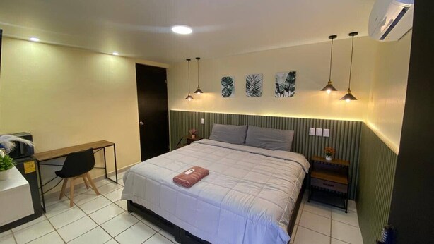 Quarto de hotel com cama, escrivaninha e cadeira. Há luminárias fixadas na parede e uma pequena mesa com uma planta.