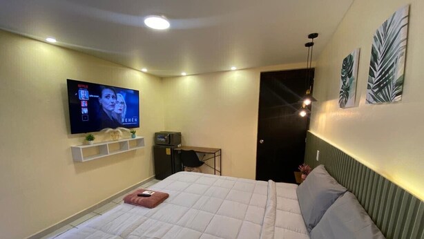 Un dormitorio con una cama, televisor montado en la pared, un escritorio con silla y una puerta.