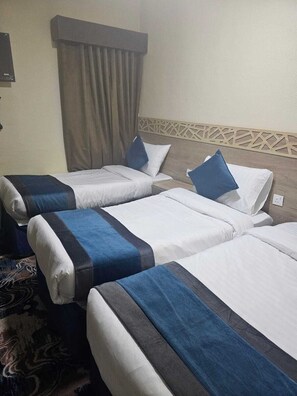 A hotel room with two beds, each with white linens and blue pillows. There is a patterned rug on the floor and a curtain on the window.