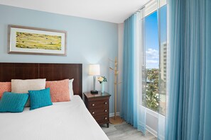 A hotel room with a large bed, a nightstand, a lamp, a framed picture, and a view of the city through a window. - Palms Resort #1603 Jr. 2BR (Destin)