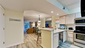 A modern kitchen with a central island, a dining area with a view, and a living room with a large window. - Sea Oats 111 (Fort Walton Beach)