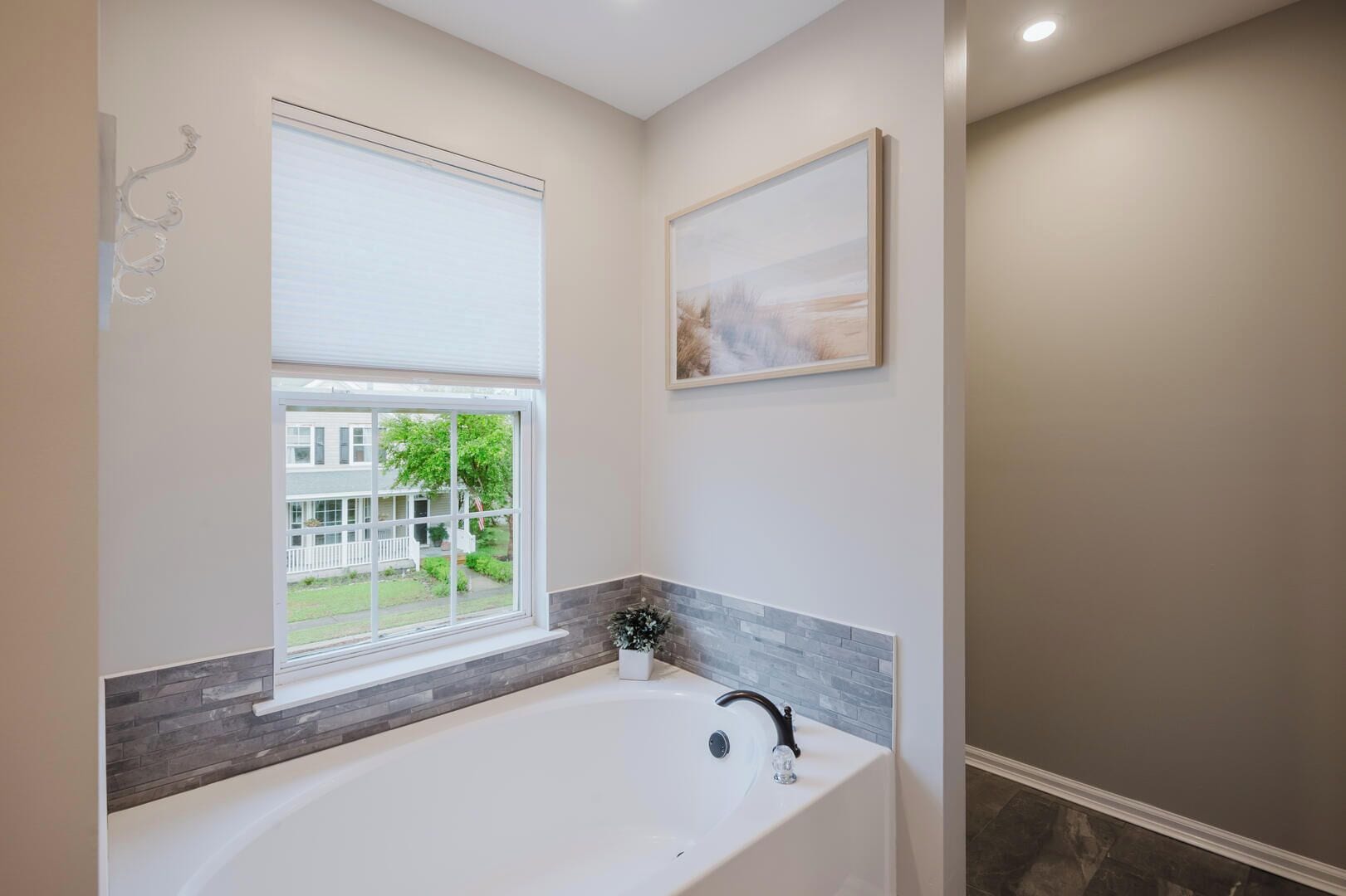 A modern bathroom with a large bathtub, a window with a view of a house, and a framed artwork on the wall.