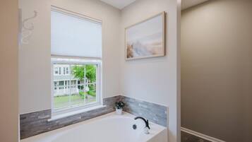 A modern bathroom with a large bathtub, a window with a view of a house, and a framed artwork on the wall.