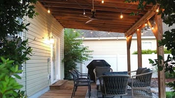 A covered patio with wooden flooring, outdoor furniture, string lights, and a grill.