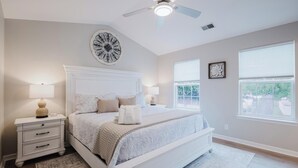 Un dormitorio con cama, mesitas de noche, ventilador de techo y dos ventanas.