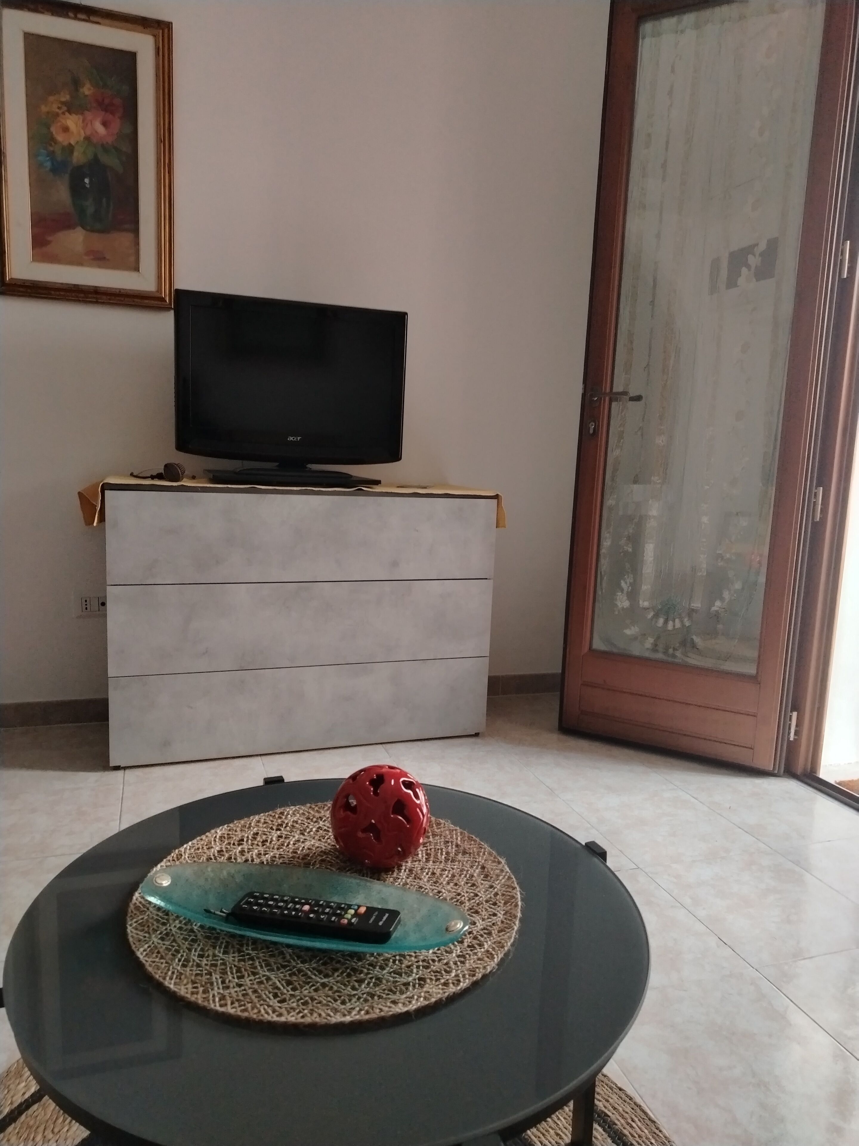 Una sala de estar con un televisor sobre un mueble bajo, una mesa de centro de vidrio con un control remoto, una pelota roja y un cuadro en la pared.