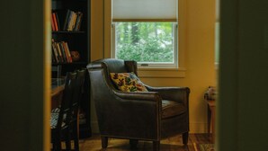 A room with a wooden floor, a leather armchair with a floral pillow, a bookshelf, and a window with blinds. - Warren Village home steps from bubbling brook (Warren)