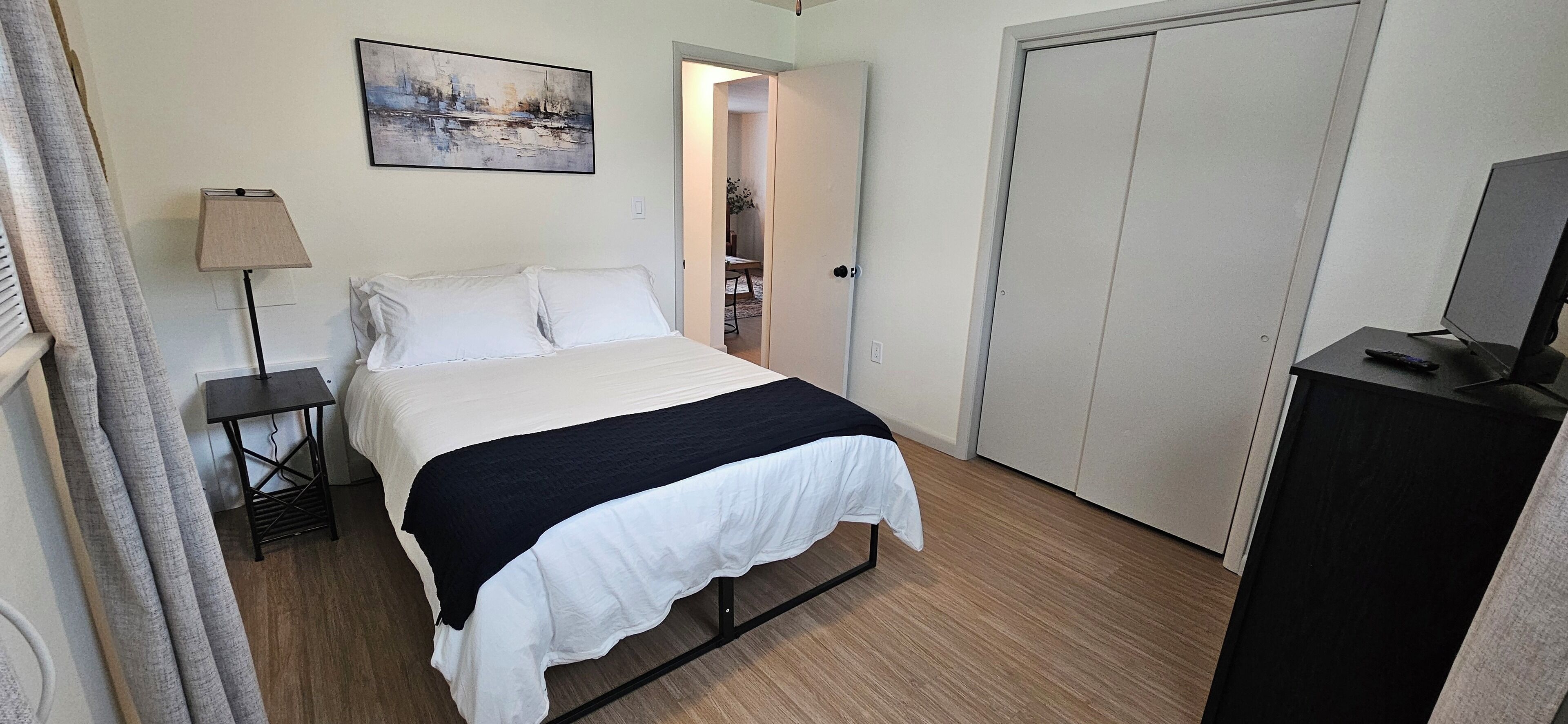 A bedroom with a bed, a bedside lamp, a painting on the wall, and a door leading to another room.