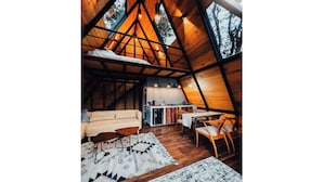 A cozy, triangular-roofed cabin interior with a sofa, dining table, and chairs, and a kitchen area visible through the open space. - Chalet Goliat by Woodland Cabins (Ciudad López Mateos)