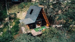 A small, A-frame wooden cabin with a sloped roof, a deck with outdoor seating, and a large window. - Chalet Goliat by Woodland Cabins (Ciudad López Mateos)
