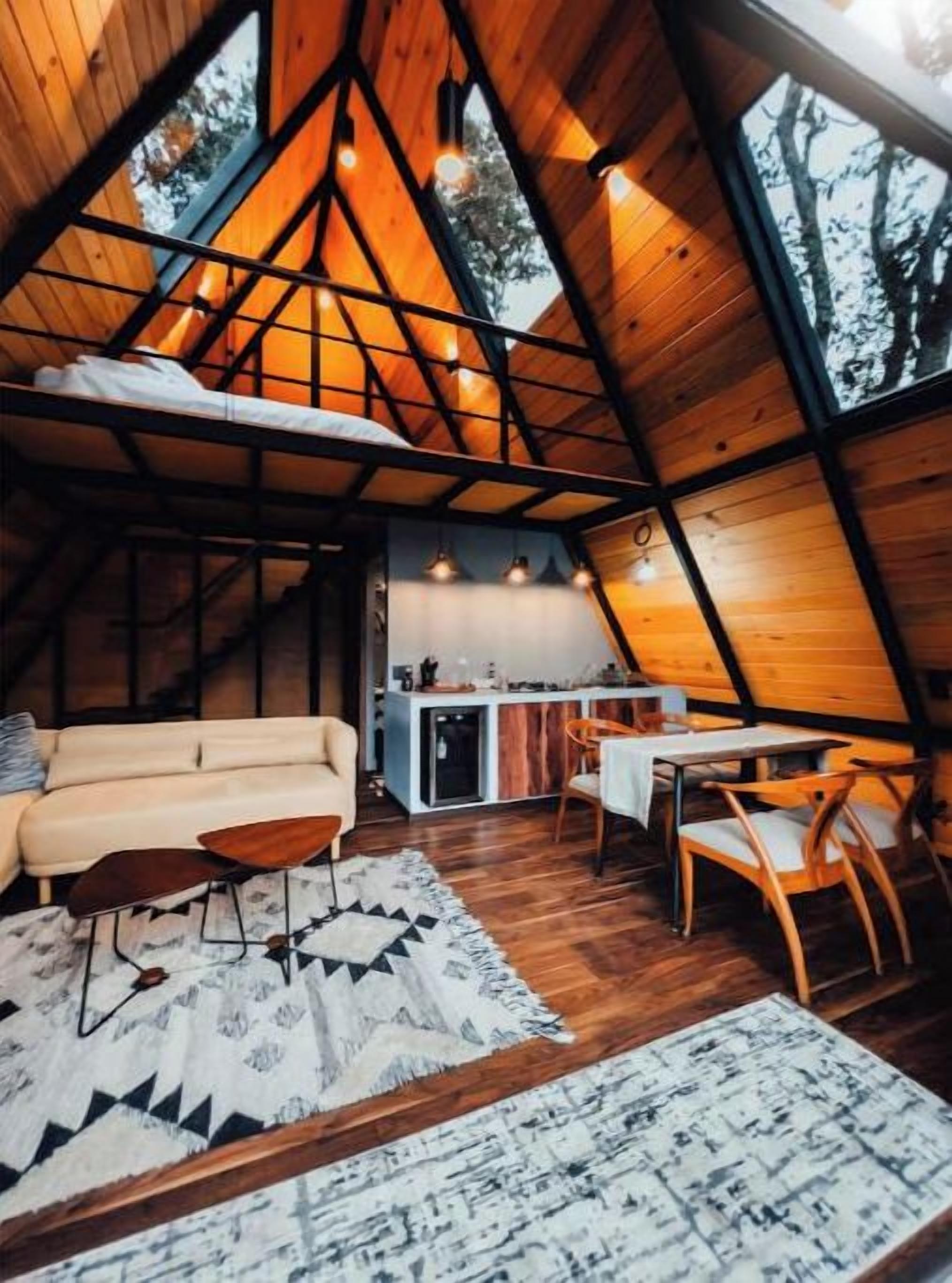 L’intérieur d’un chalet accueillant, doté d’un toit triangulaire, comprend un canapé, une table à manger et des chaises, et une cuisine ouverte sur le reste de l’espace.
