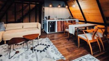L’intérieur d’un chalet accueillant, doté d’un toit triangulaire, comprend un canapé, une table à manger et des chaises, et une cuisine ouverte sur le reste de l’espace.