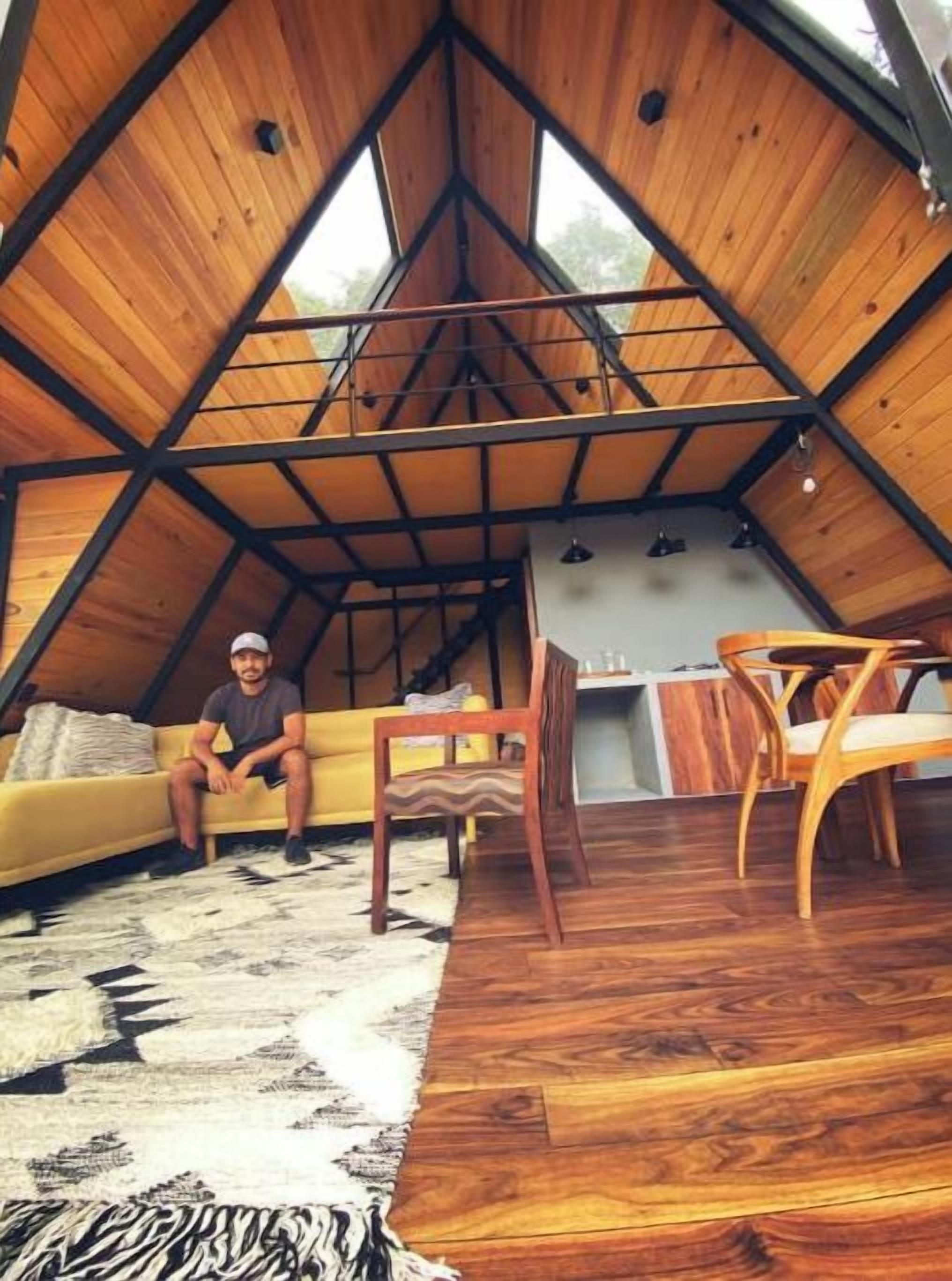 Ein gemütliches Dachzimmer mit schrägverdeckter Holzbalkendecke, einem gelben Sofa, einem Holzesstisch und einem gemusterten Teppich.