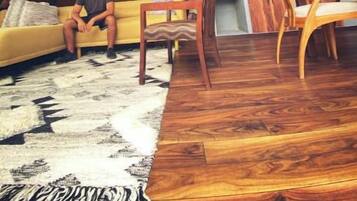 Ein gemütliches Dachzimmer mit schrägverdeckter Holzbalkendecke, einem gelben Sofa, einem Holzesstisch und einem gemusterten Teppich.