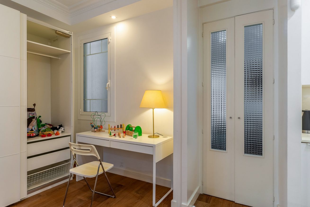 A room with a white desk, a chair, a lamp, a mirror, and a closet with a window.