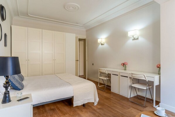 A bedroom with a bed, bedside table, lamp, desk, chairs, and a small table with a cup.