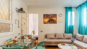 A modern living room with a glass dining table, teal chairs, a beige sofa, and a painting of a sunset.