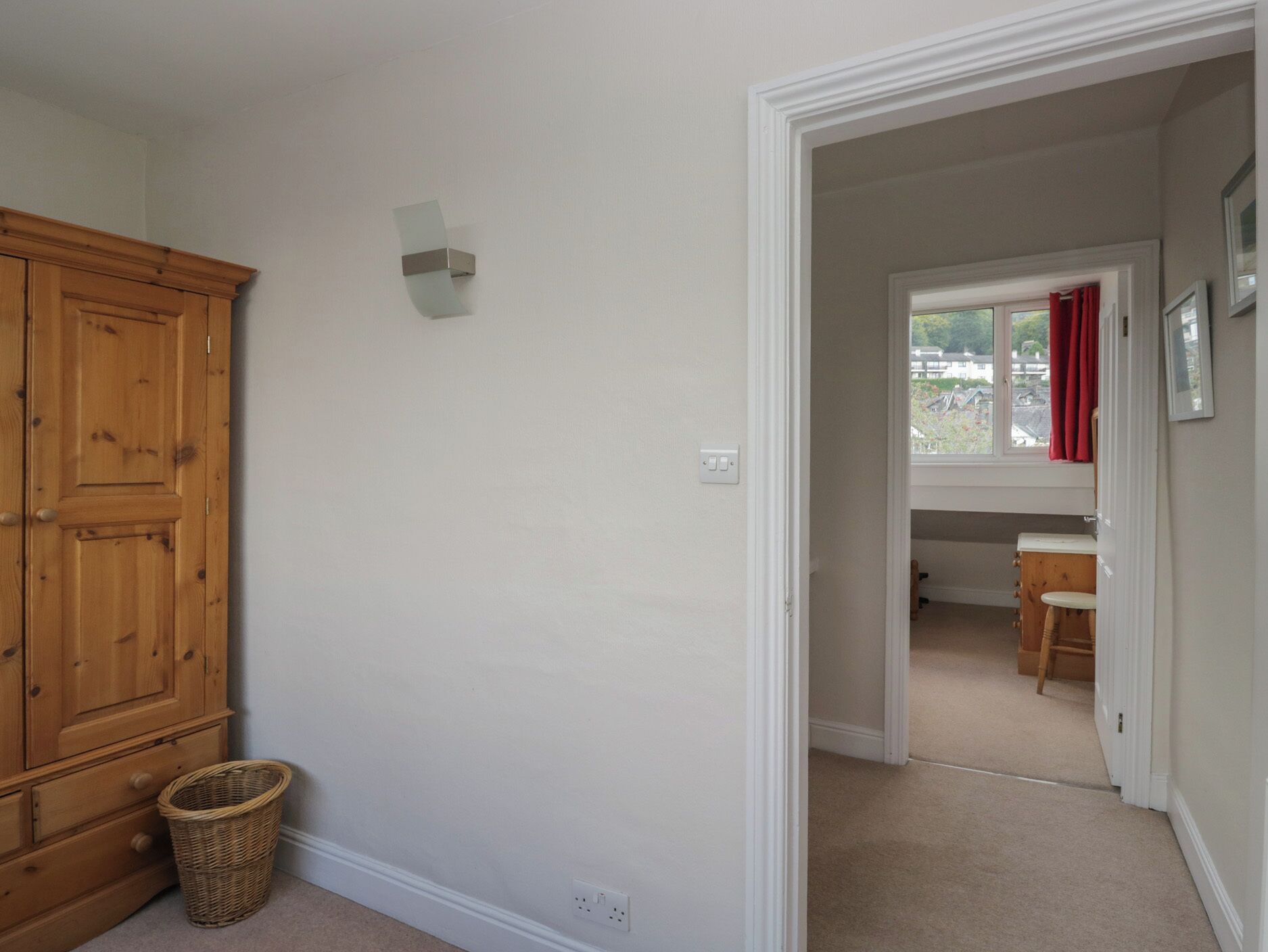 A room with a wooden wardrobe, a small basket, and a window with red curtains.