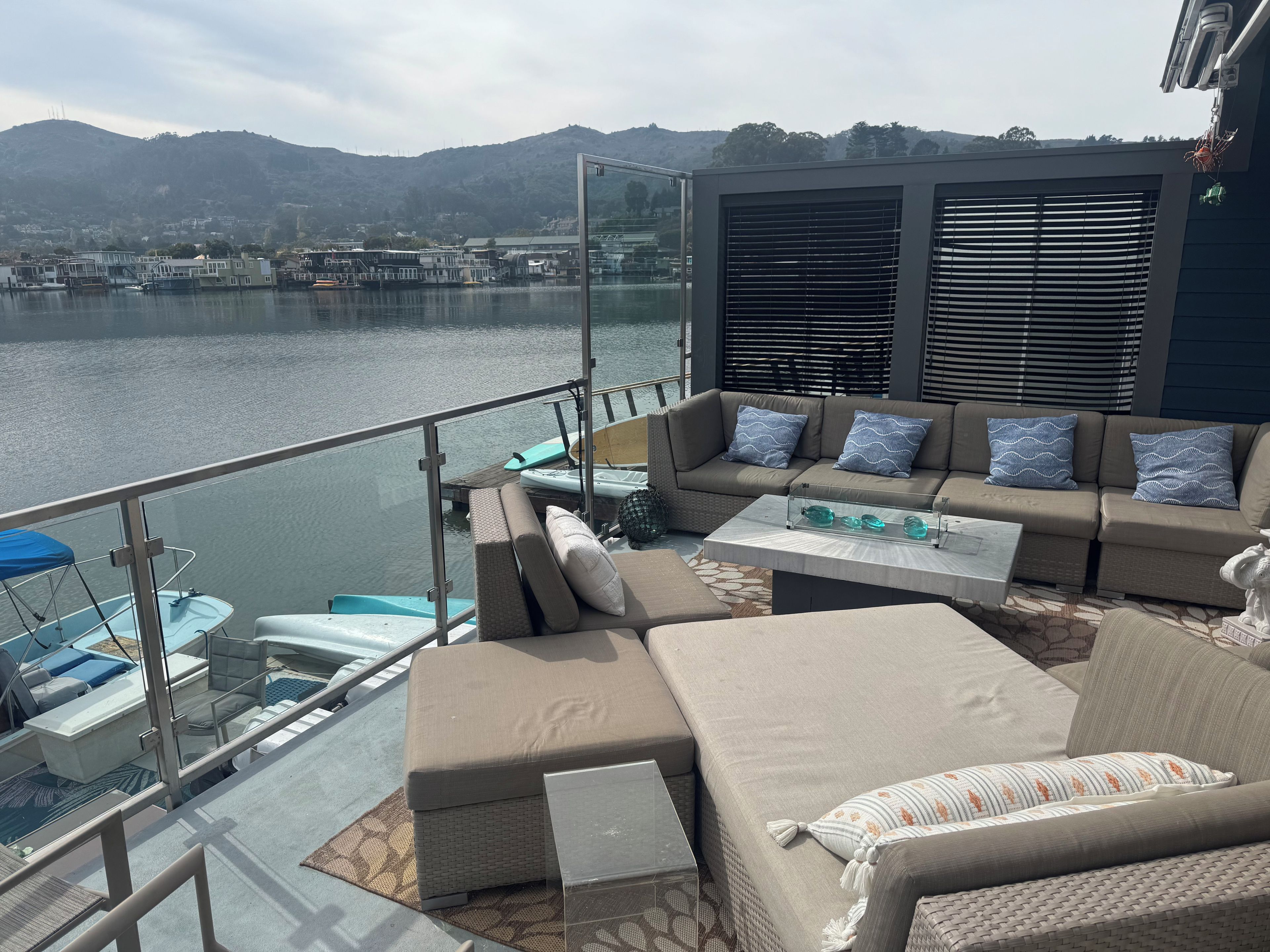 Eine Terrasse mit Gartenmöbeln, einer Glasreling und Blick auf ein Gewässer mit Gebäuden im Hintergrund.