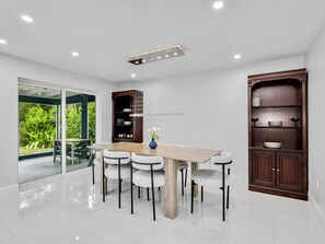 A modern dining area with a wooden table and chairs, a glass door leading to a garden, and a dark wooden cabinet with shelves. - 1.5 miles 5 min 🚗 to beaches, King Beds, BBQ, Nintendo Switch 🎮, Pet friendly 🐶. (Naples)