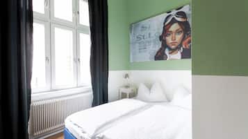 Un dormitorio con cama, una ventana con cortinas, un radiador, una mesita de noche con lámpara y un cartel en la pared.