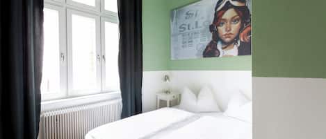 Un dormitorio con cama, una ventana con cortinas, un radiador, una mesita de noche con lámpara y un cartel en la pared.