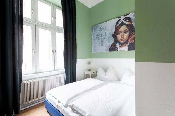A bedroom with a bed, a window with curtains, a radiator, a nightstand with a lamp, and a wall with a poster.