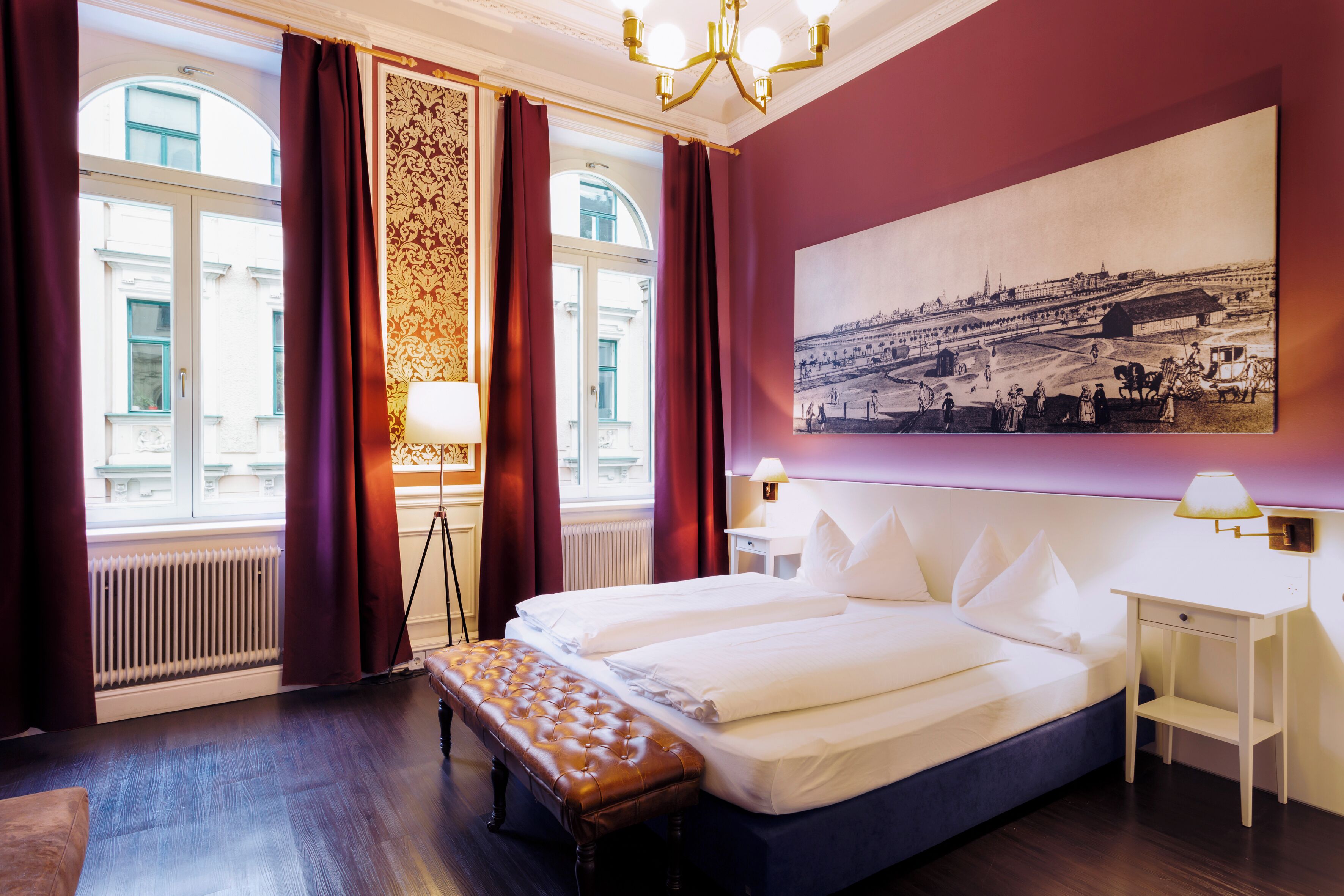 A hotel room with a large bed, a bench, a nightstand, a lamp, a window with curtains, and a wall-mounted picture.