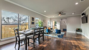 Un vaste salon doté de planchers en bois franc, un coin repas avec une table et des chaises, une grande fenêtre donnant sur un jardin, et un ventilateur de plafond.
