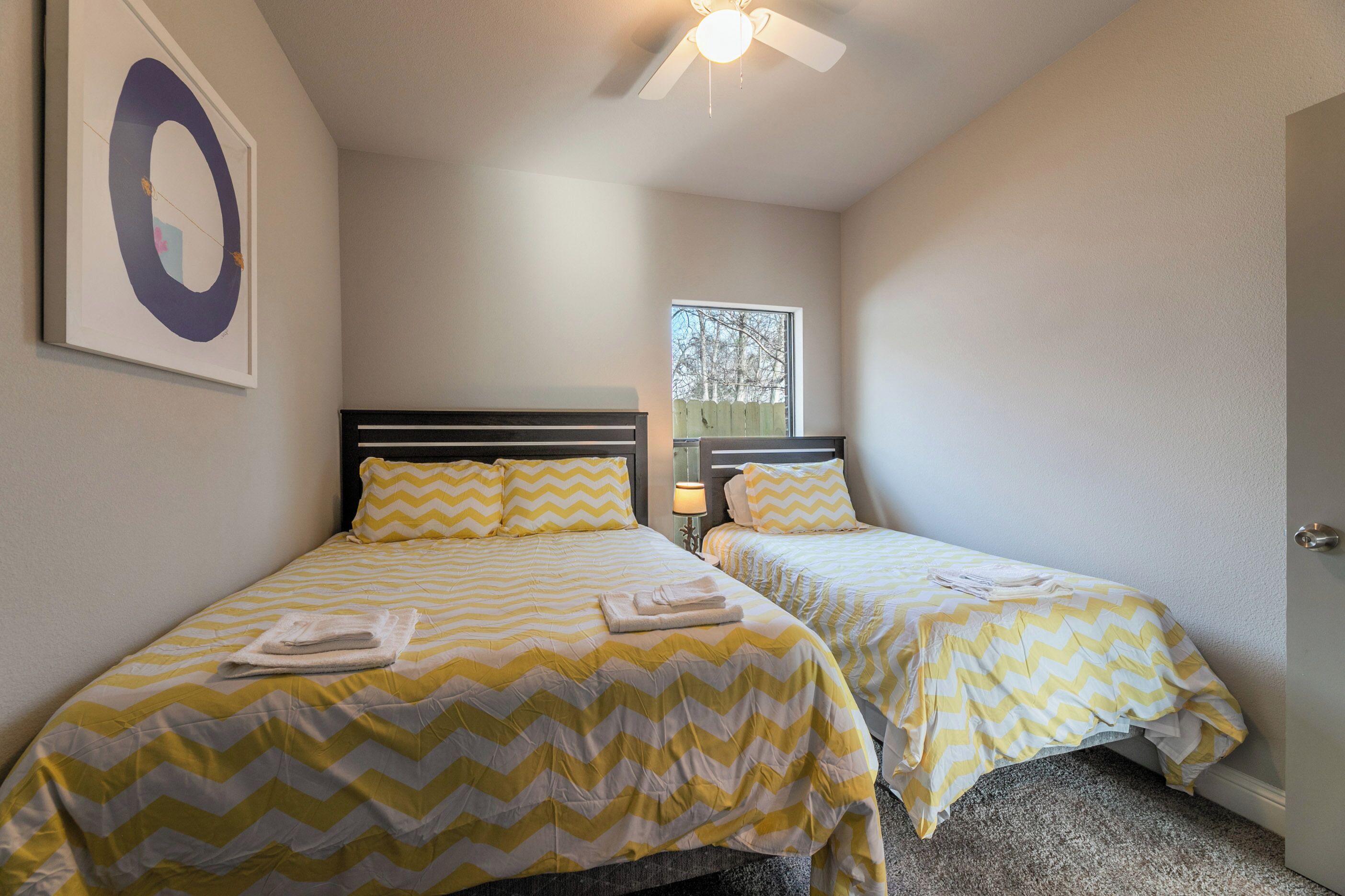 A hotel room with two beds, each with a yellow and white chevron-patterned bedspread, a ceiling fan, and a window with a view of trees.