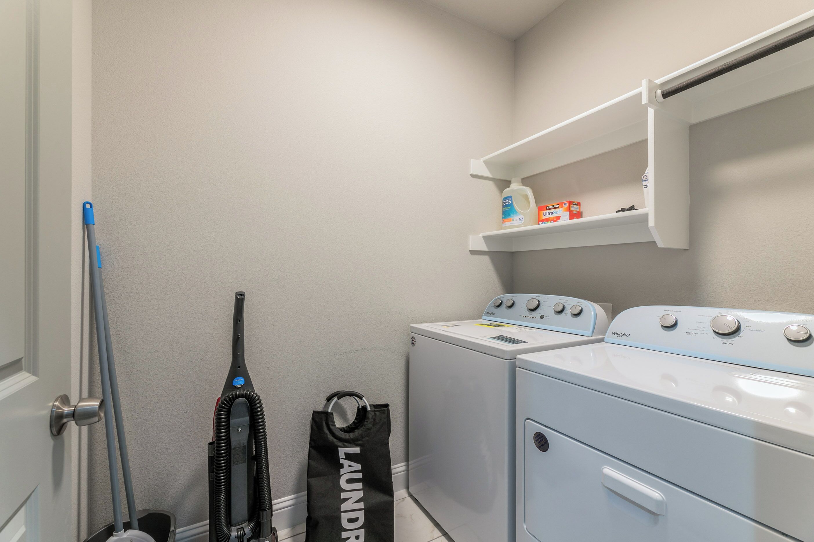 A laundry area with a washing machine, a dryer, a shelf with detergent, and a vacuum cleaner.