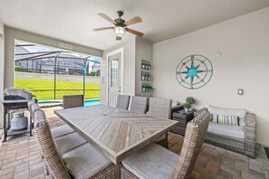An outdoor patio with a wooden table and wicker chairs, a ceiling fan, a grill, and a view of a green lawn and a greenhouse. - 5BR Pool Home, Near Disney, Orlando (Kissimmee)