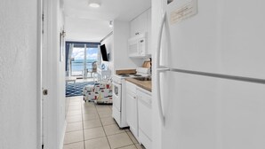 A narrow hallway leading to a kitchen with white cabinets, a microwave, and a refrigerator. There is a blue patterned rug on the floor and a view of the sea through a glass door.