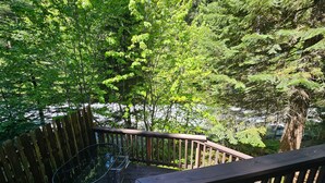 A wooden deck with a metal table and chairs, surrounded by lush green trees and a view of a body of water.